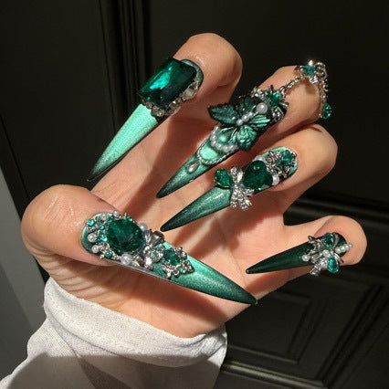 Handmade Emerald Green Cat Eye Long Stiletto Press On Nails. Luxury witchy false nails decorated with 3D green butterfly charms, large emerald crystals, pearls, silver rhinestones and dangling gem chains. Reusable fake nails for parties and cosplay.